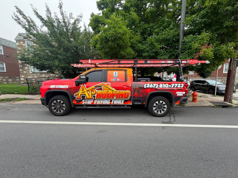 The red AP Roofing truck at the curb with ladders on the rack